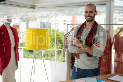Portrait of smiling male designer with arms crossed standing by clothes rack