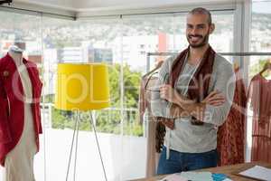 Portrait of smiling male designer with arms crossed standing by clothes rack