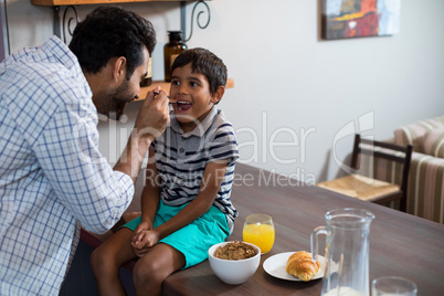 Happy father feeding cereal breakfast to son sitting on table