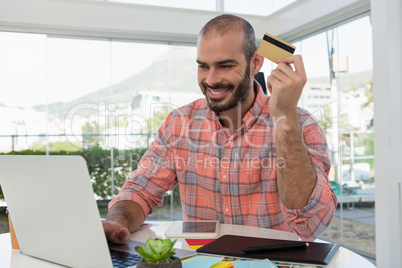 Smiling graphic designer holding credit card while using laptop at desk