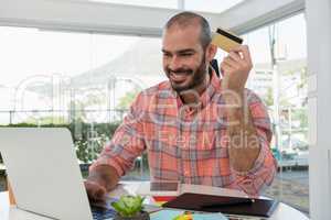 Smiling graphic designer holding credit card while using laptop at desk