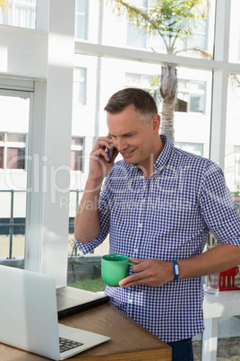 Designer holding cup talking on mobile phone while looking at laptop