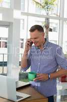 Designer holding cup talking on mobile phone while looking at laptop