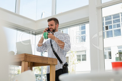 Businessman having drink while talking on mobile phone while standing at desk