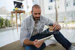 Designer holding disposable cup using tablet computer while sitting on floor