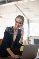 Portrait of woman talking on phone while using laptop in office