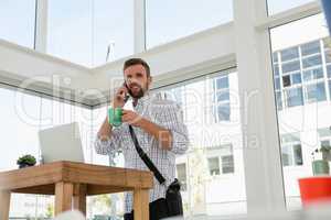 Businessman having drink while talking on phone while standing at desk
