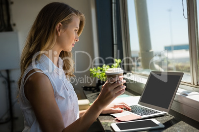 Businesswoman holding coffee cup while using laptop