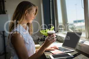 Businesswoman holding coffee cup while using laptop
