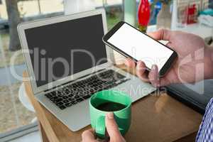 Cropped hand of designer using smart phone by laptop