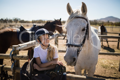Smiling girl leaning on the fence and looking at the white horse in ranch
