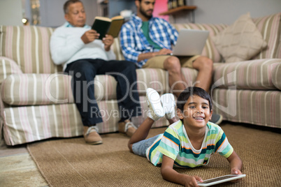 Smiling boy using tablet while lying on carpet with father and grandfather in background