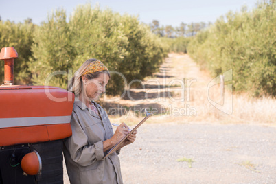 Woman leaning on tractor while writing on clipboard