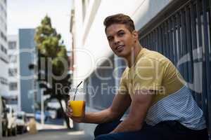 Man holding juice looking away while sitting by wall in city