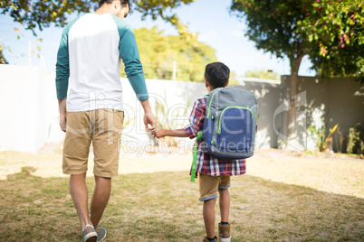 Rear view of father holding hand of son with backpack in yard