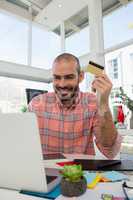 Smiling designer holding credit card while using laptop at desk