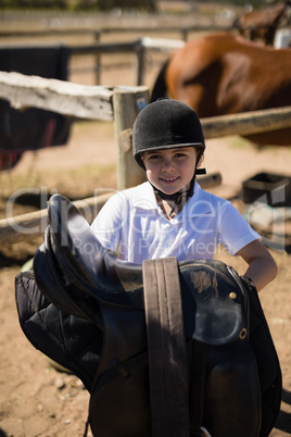 Smiling girl holding horse saddle in the ranch