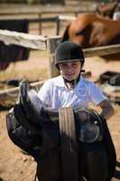Smiling girl holding horse saddle in the ranch