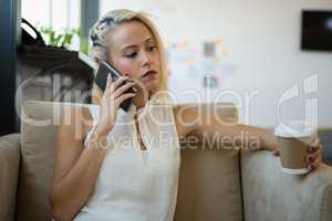 Woman with coffee cup talking on phone at sofa in office