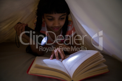 Girl holding flashlight while reading novel under blanket