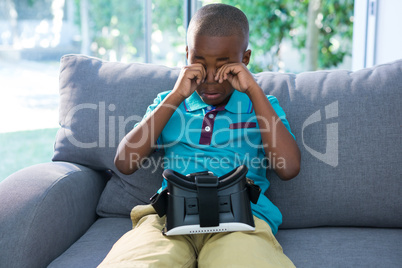 Boy rubbing eyes while sitting with VR headset on sofa