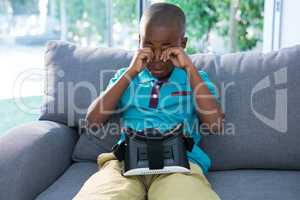 Boy rubbing eyes while sitting with VR headset on sofa