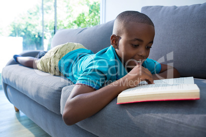 Smiling boy reading novel while lying on sofa at home