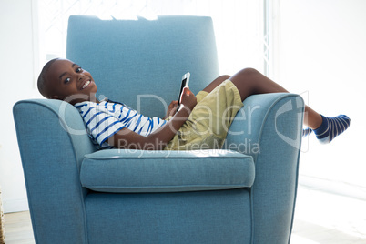 Portrait of smiling boy with mobile phone while lying on armchair at home