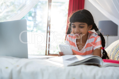 Girl using mobile phone while lying by book and laptop on bed