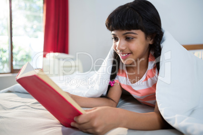Smiling girl reading novel on bed at home