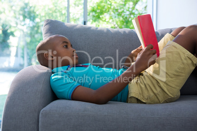 Side view of boy lying while reading novel on sofa at home