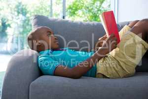 Side view of boy lying while reading novel on sofa at home