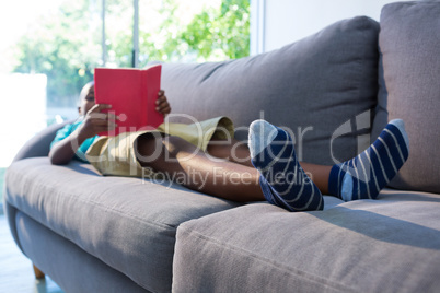 Boy lying on sofa while reading novel against window at home