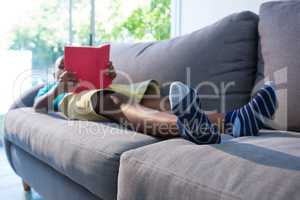 Boy lying on sofa while reading novel against window at home