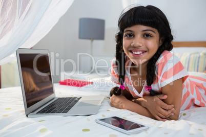 Portrait of smiling girl lying with mobile phone and laptop on bed