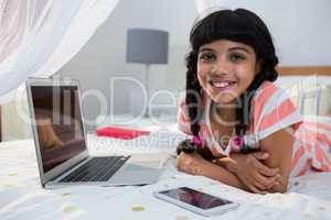 Portrait of smiling girl lying with mobile phone and laptop on bed