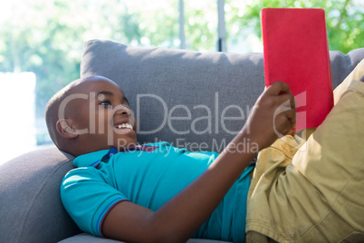 Side view of smiling boy lying while reading novel on sofa at home