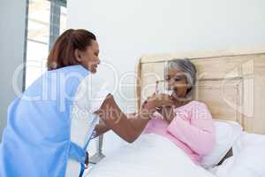 Female doctor giving glass of water to sick senior woman on bed