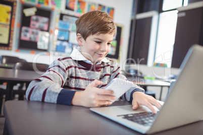 Smiling schoolboy using laptop and mobile phone on desk at classroom