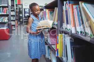 Girl reading book by shelf in library