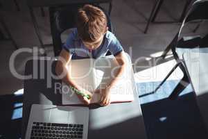 Boy writing in book by laptop at desk