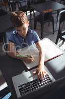 Boy using laptop while writing in book at school