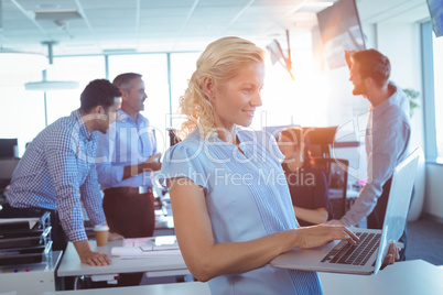 Businesswoman working on laptop with team discussing in background