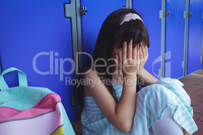 Schoolgirl covering face with hands while sitting by lockers