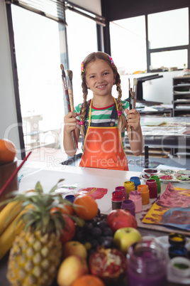 Portrait of smiling girl holding paintbrushes by desk