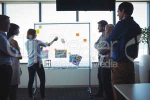 Businesswoman sticking adhesive notes on whiteboard while colleagues standing at office