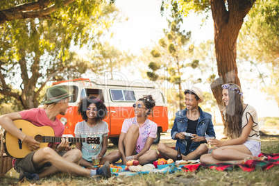 Smiling friends looking at man playing guitar while sitting on field