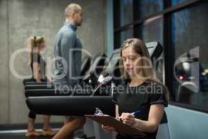 Woman writing on clipboard with people exercising on treadmill
