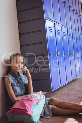 Smiling elementary schoolgirl talking on mobile phone while sitting by lockers