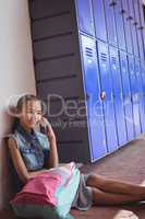 Smiling elementary schoolgirl talking on mobile phone while sitting by lockers